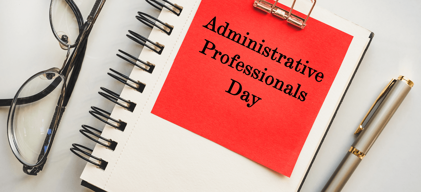 Notebook with a red note reading Administrative Professionals Day beside reading glasses and a pen