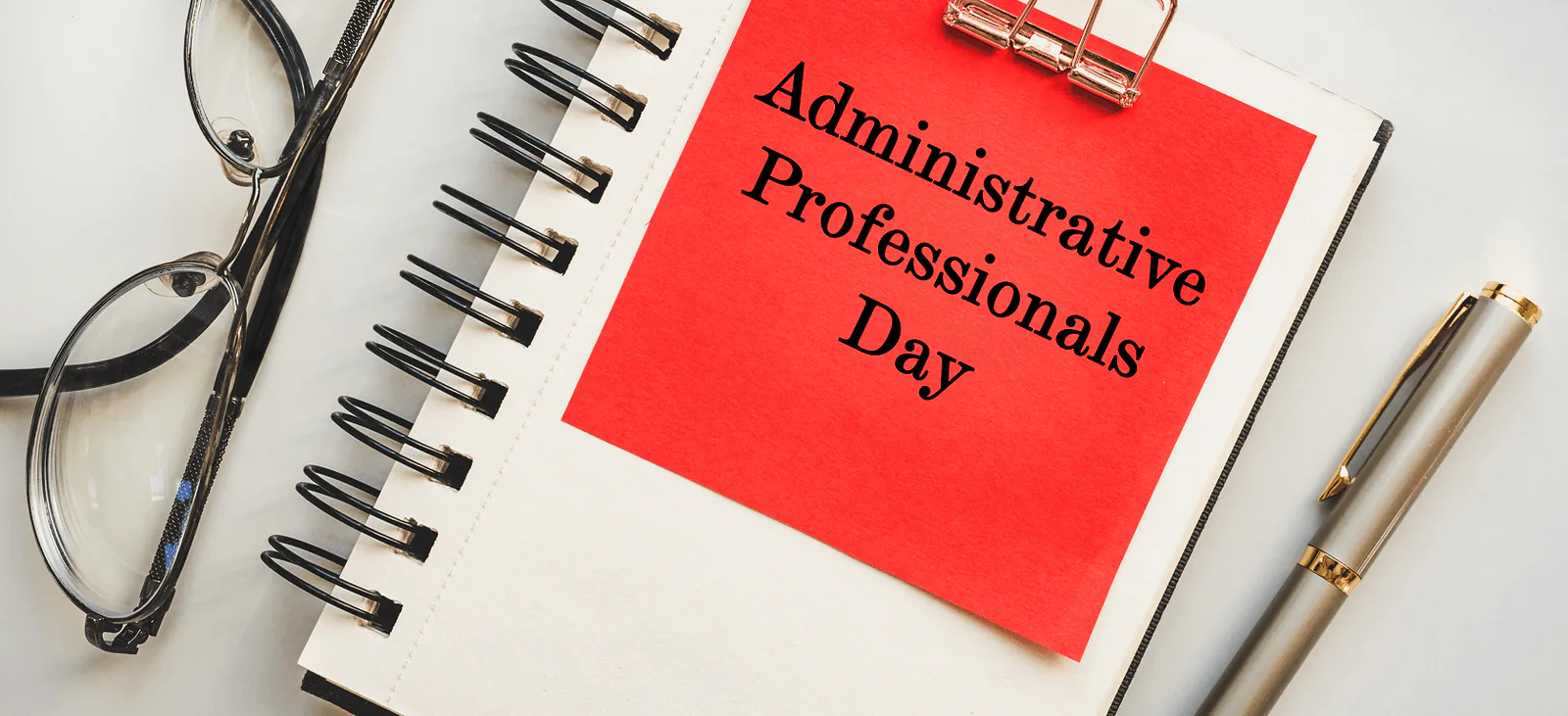 Notebook with a red note reading Administrative Professionals Day beside reading glasses and a pen