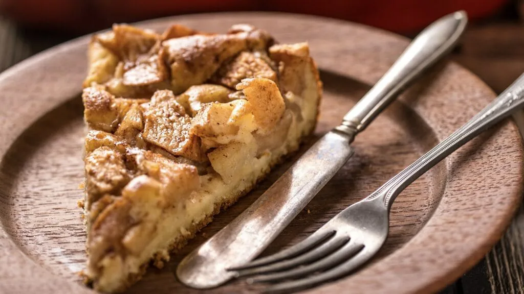 Slice of cinnamon apple pie on a wooden plate with an antique fork and knife