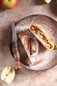 Two slices of apple strudel dusted with powdered sugar on a plate beside fresh apples
