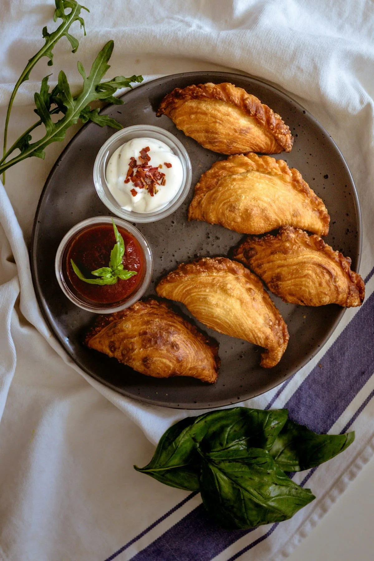 Baked empanadas on a plate with sour cream and tomato dipping sauces, basil and arugula alongside