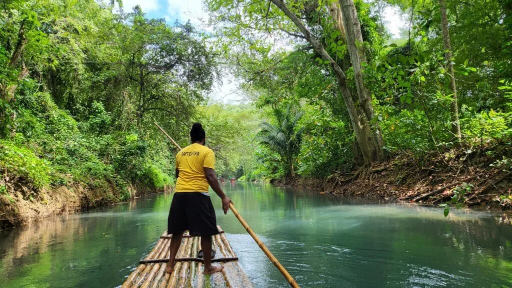 A guide poles a bamboo raft down a calm river through dense tropical forest