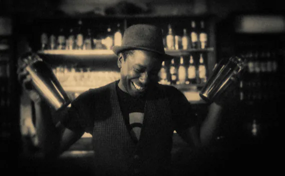 Bartender laughing while holding up two cocktail shakers in front of a stocked back bar