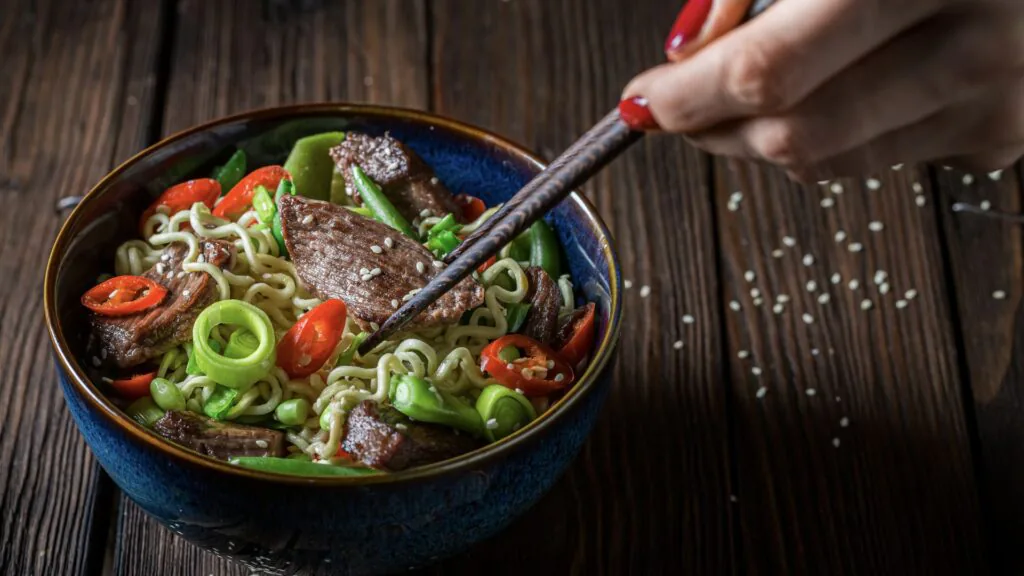 Bowl of noodles with sliced beef, chili and spring onion being lifted with chopsticks