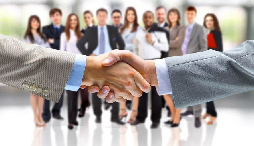 Two people in business suits shaking hands with a group of colleagues blurred behind them