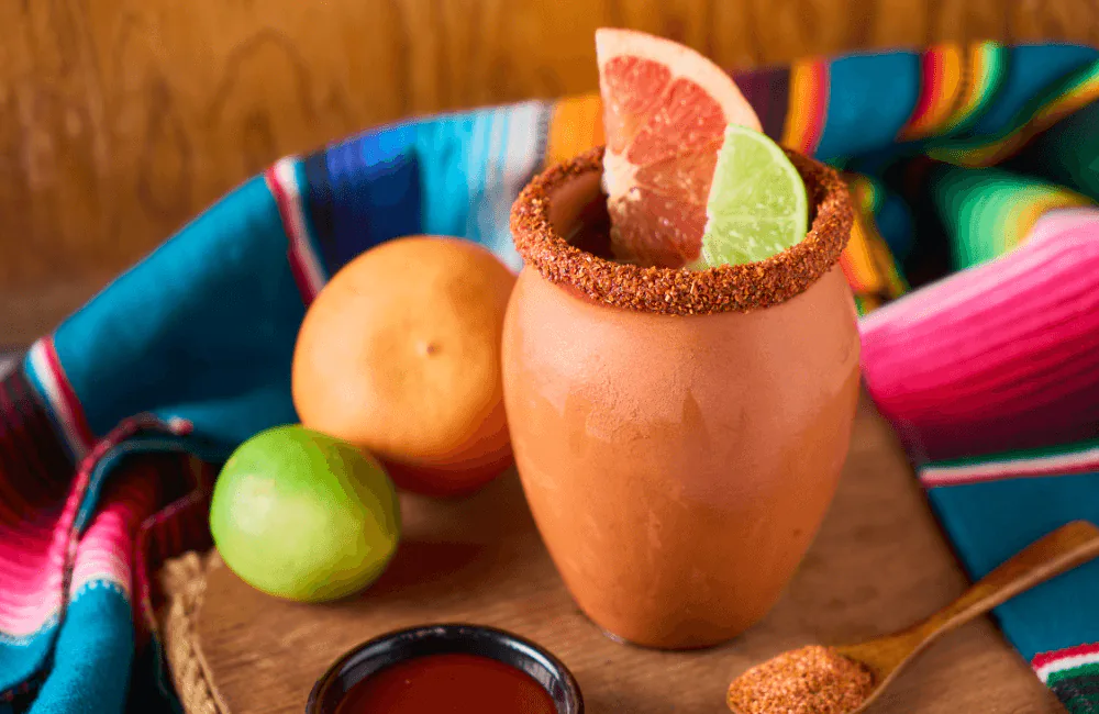 Cantarito cocktail in a clay cup with a chili salt rim, grapefruit and lime wedges on a serape