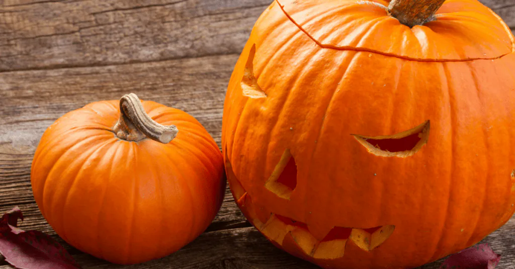 A carved jack-o’-lantern beside a small uncarved pumpkin on a weathered wood table