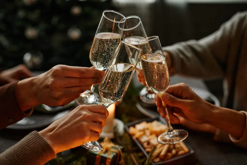 Four hands clinking champagne flutes at a holiday dinner table with a lit Christmas tree behind