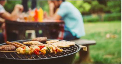 Skewers and burgers cooking on a charcoal grill at a backyard barbecue with friends behind