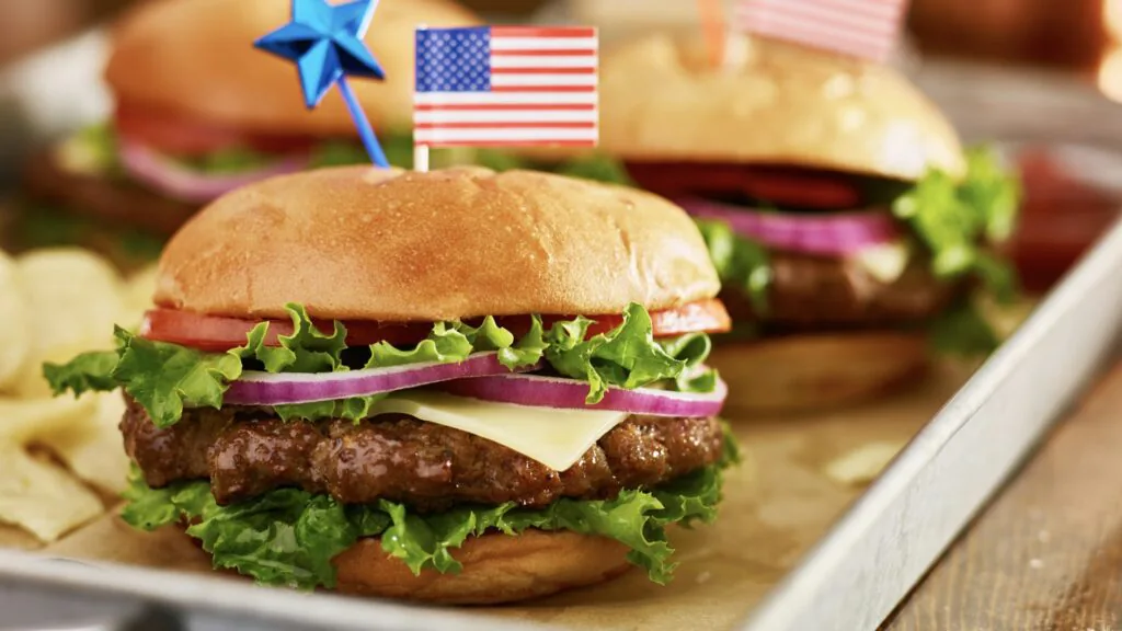 Cheeseburgers with lettuce, tomato and red onion topped with small American flag picks beside chips