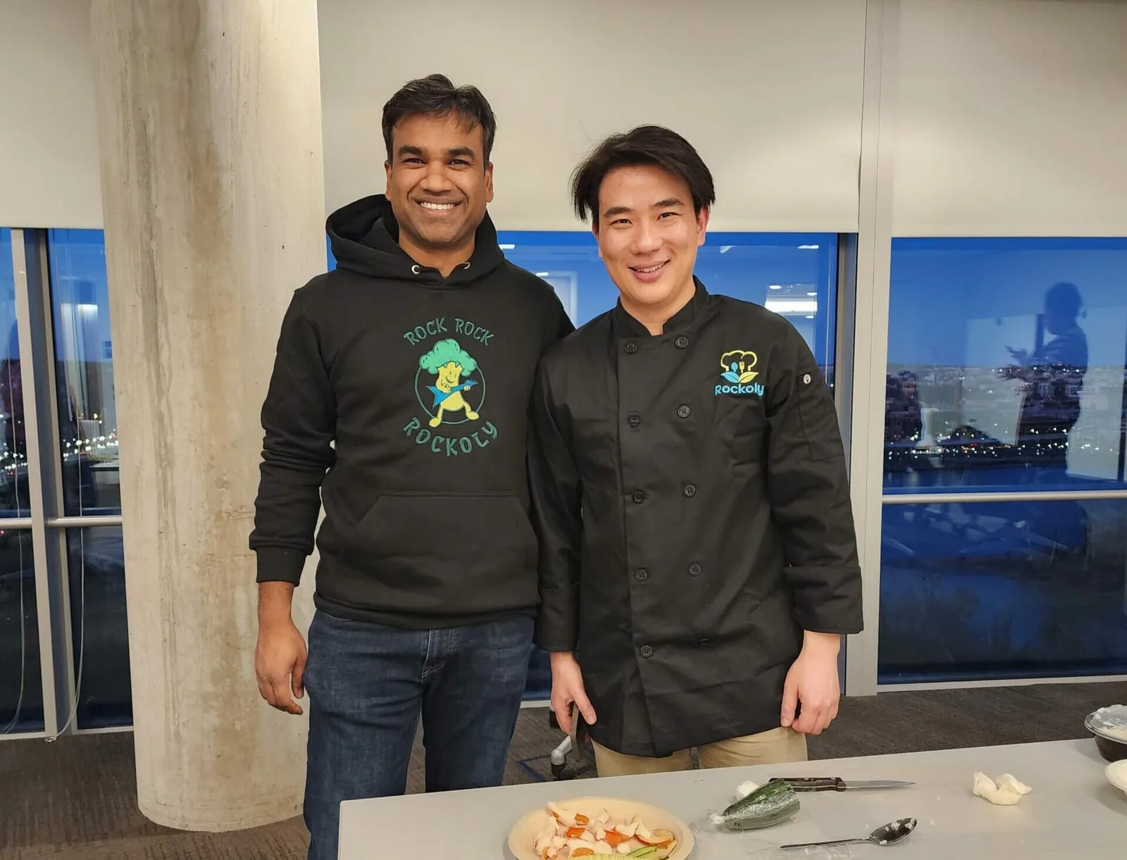 A chef in a Rockoly jacket and a guest in a Rockoly hoodie smiling beside a plated dish at an office event