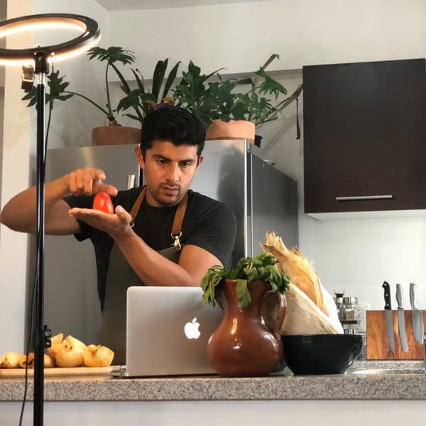 Chef Arturo holding a tomato in front of a ring light and laptop while filming a virtual cooking class
