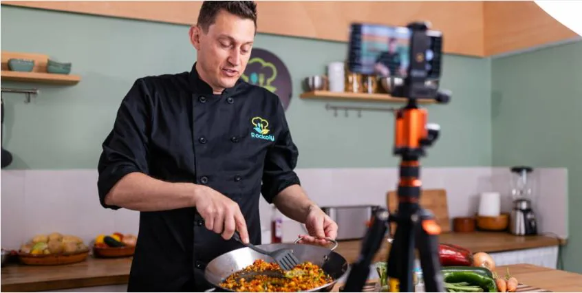 Chef in a black chef's jacket cooking a skillet dish while a phone on a tripod films the class