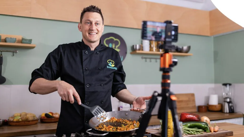 Chef in a black jacket cooking a skillet dish to camera with a phone mounted on a tripod