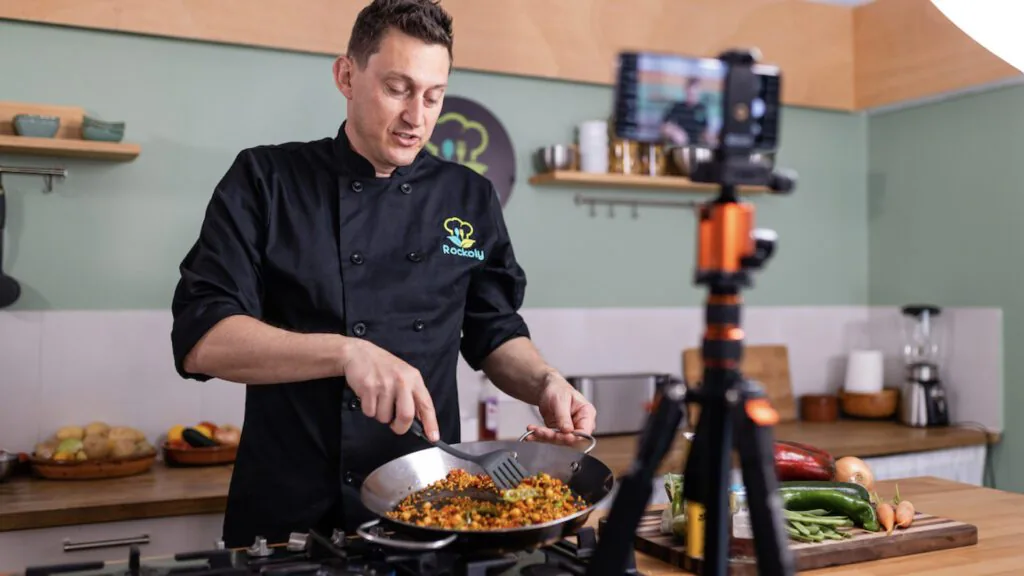 Chef Josh cooking paella in a pan while filming the class on a phone mounted on a tripod