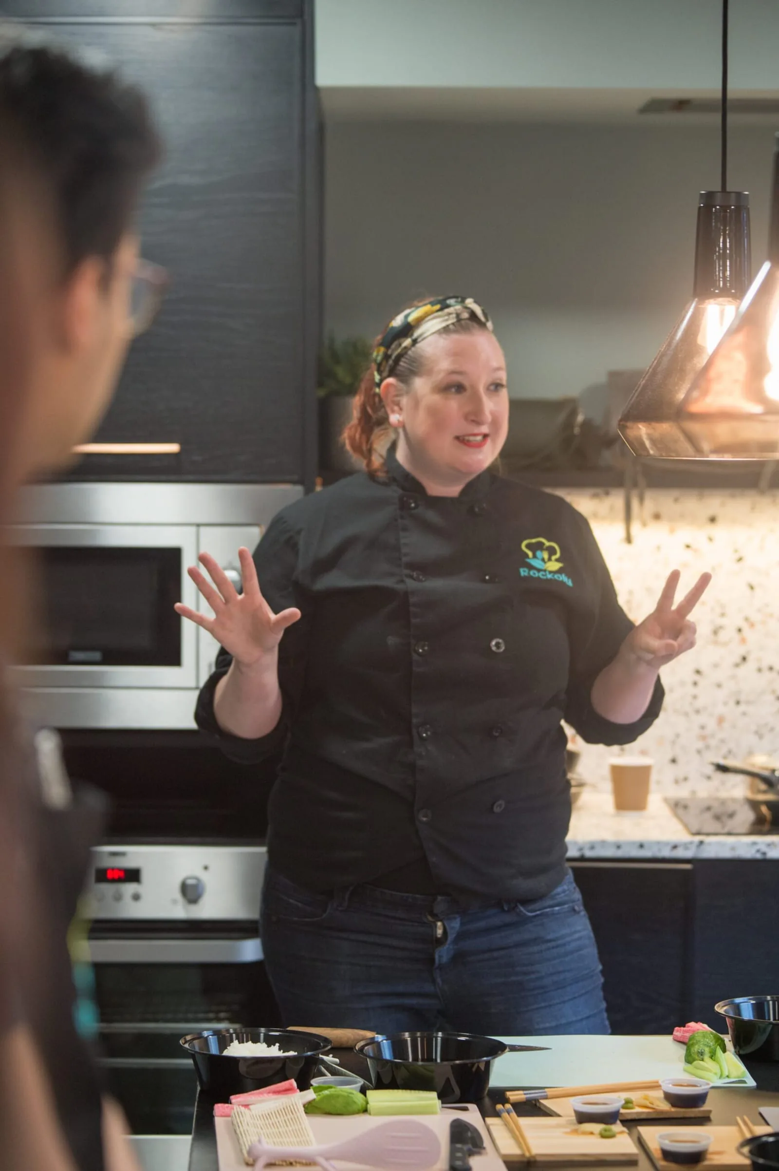 Chef in a black chef's coat gesturing while leading a hands-on cooking class in a home kitchen