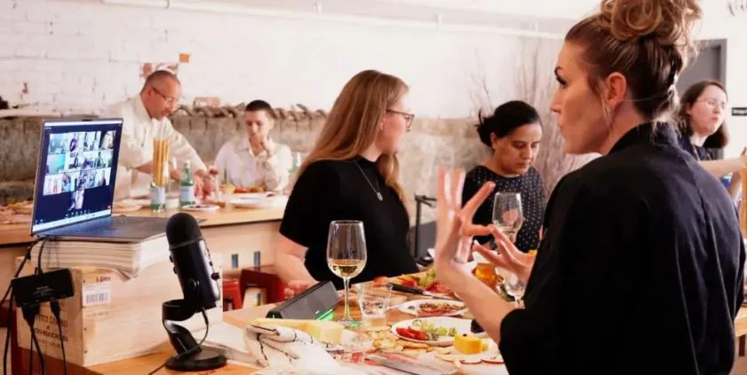 Chef talking to guests around a table of food and wine while a laptop streams the class to remote colleagues