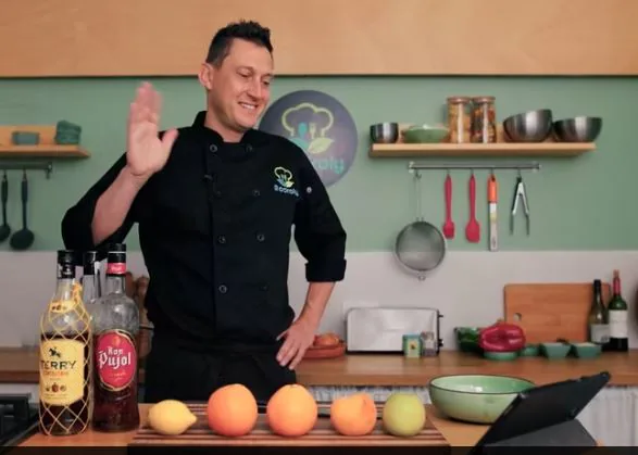 Chef waving at the camera in a studio kitchen beside rum bottles and citrus for a virtual class