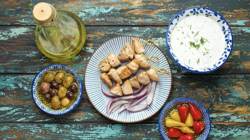 Chicken souvlaki skewers over sliced red onion with bowls of tzatziki, olives, peppers and olive oil