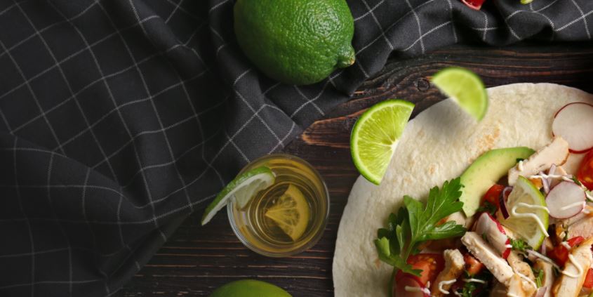 Chicken taco with avocado, radish and cilantro beside lime wedges and a shot of tequila