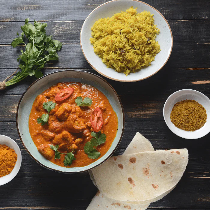 Overhead view of chicken tikka masala with yellow rice, chapati and bowls of spices on dark wood