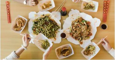 Overhead view of hands with chopsticks reaching for takeout noodles and dumplings spread across a table