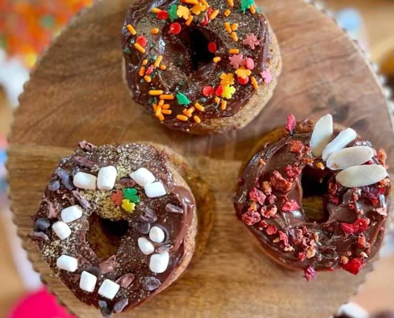 Three chocolate-glazed baked donuts topped with sprinkles, mini marshmallows and berries on a wooden board