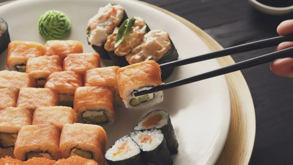 Chopsticks lifting a salmon-topped sushi roll from a plate of assorted maki with wasabi