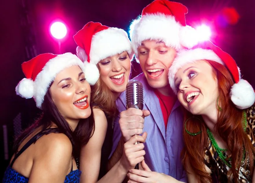 Four friends in Santa hats singing into a shared microphone under pink party lights