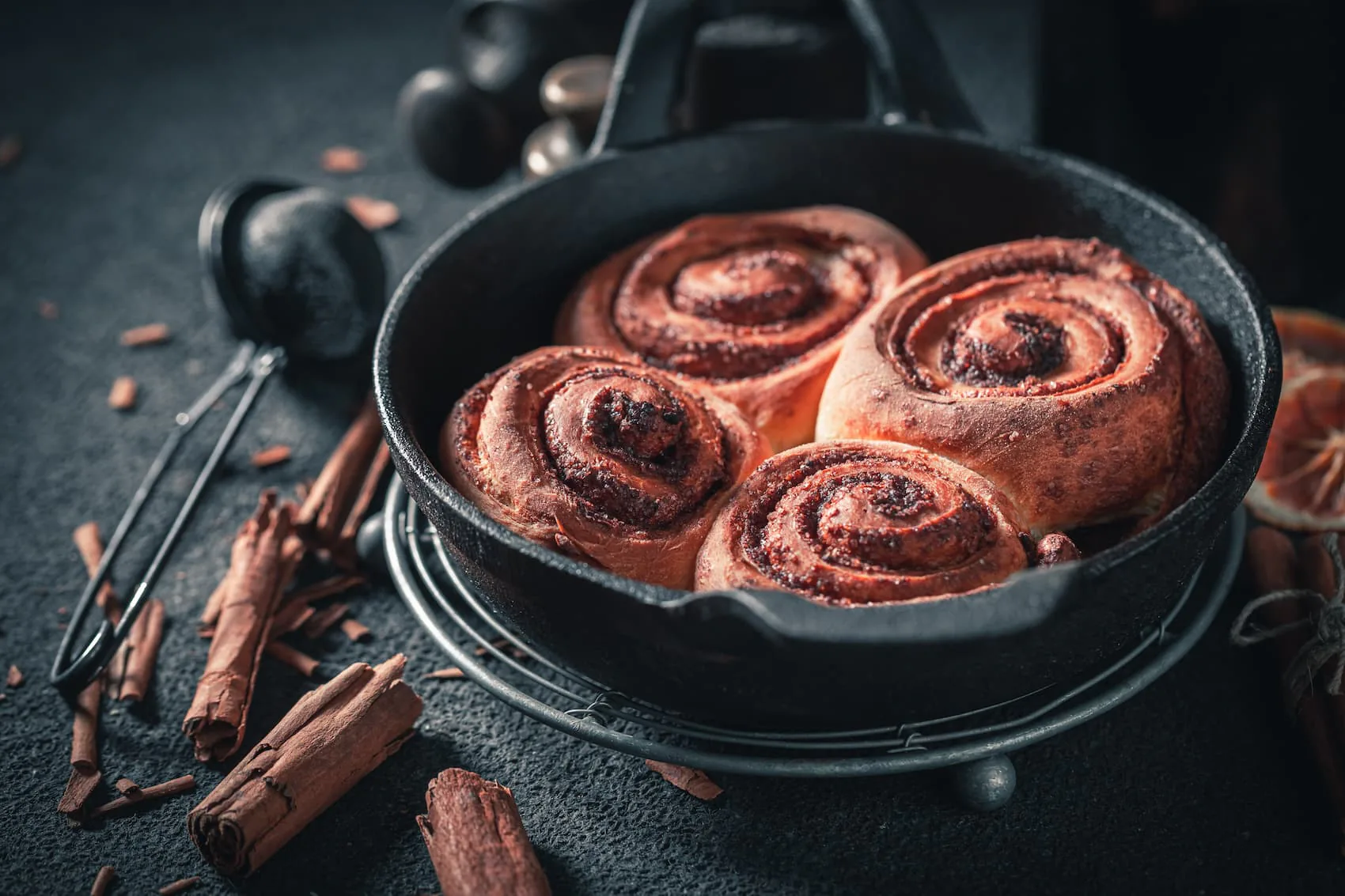 Freshly baked cinnamon rolls in a cast iron pan on a dark table with scattered cinnamon sticks