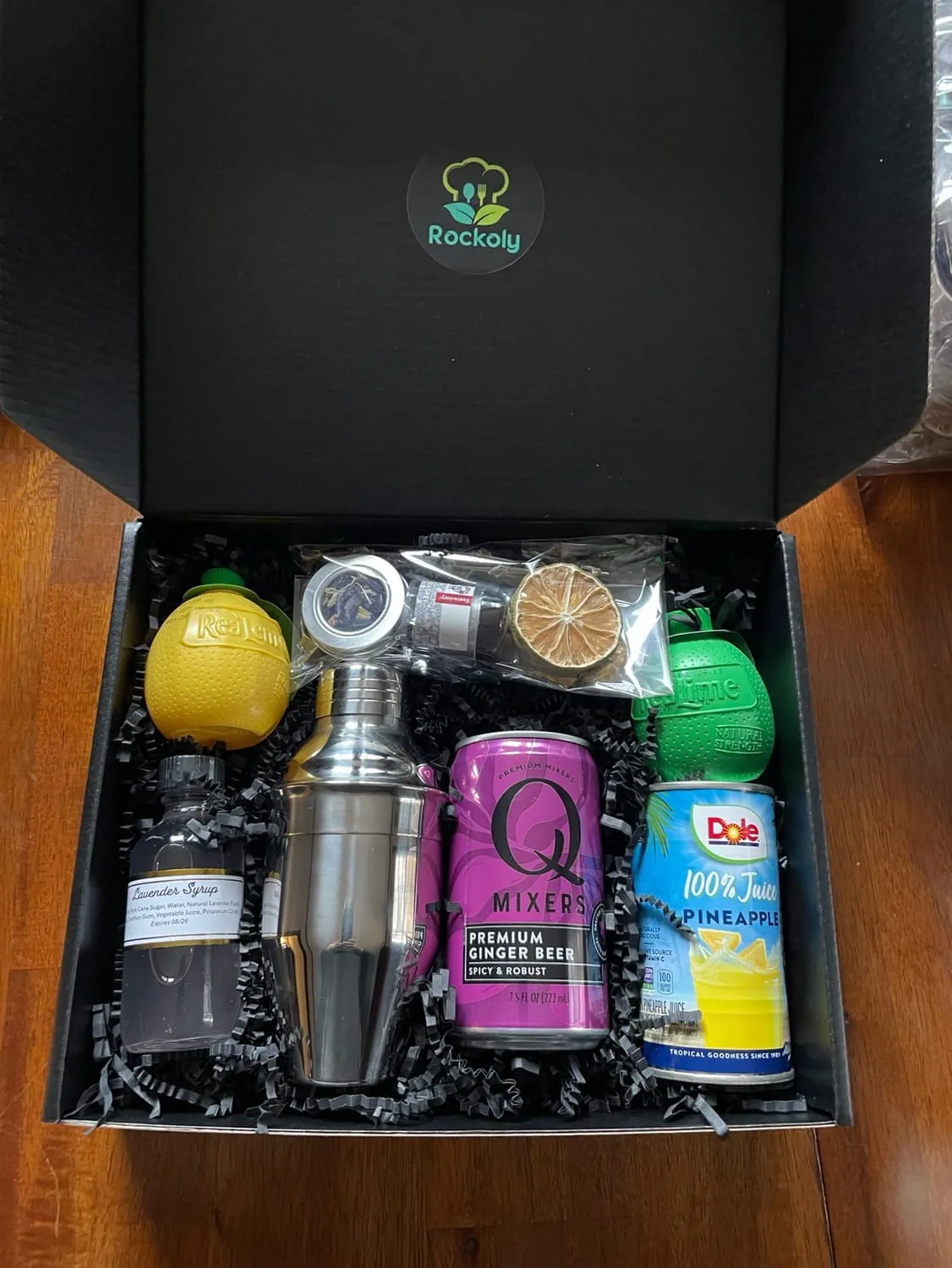 Open cocktail kit box with a steel shaker, ginger beer, pineapple juice, bitters, lemon and dried lime