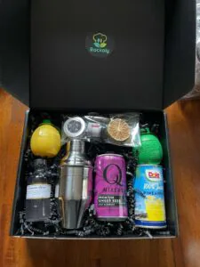 Cocktail kit box holding a steel shaker, ginger beer, pineapple juice, bitters and citrus