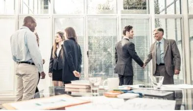 Two colleagues shaking hands in a bright glass-walled meeting room as their team looks on