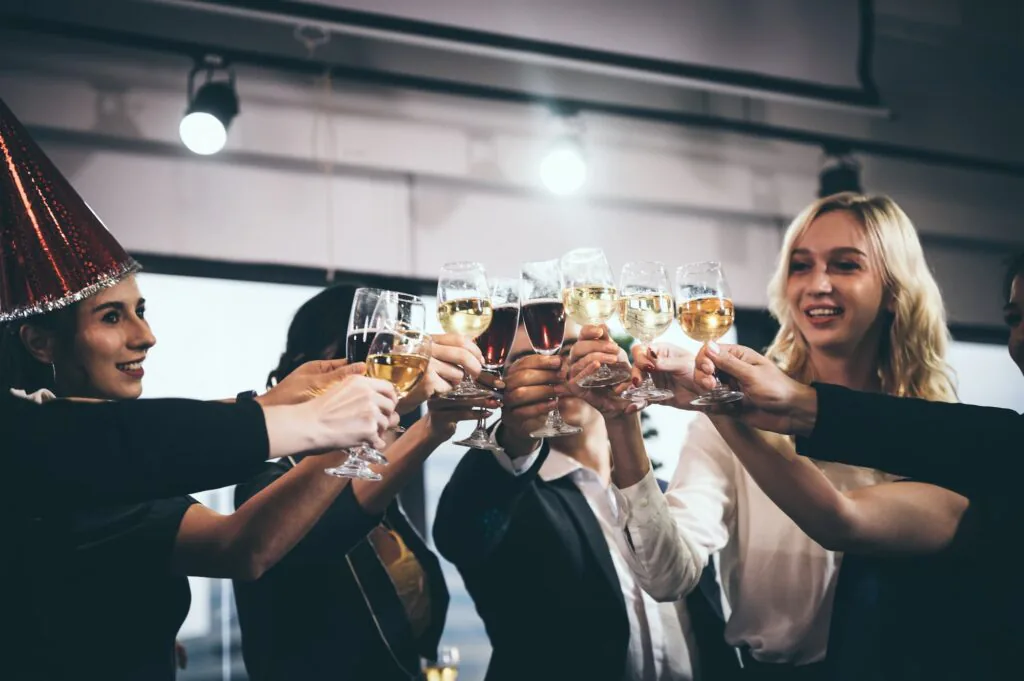 Group of colleagues raising champagne and wine glasses in a toast, one wearing a party hat