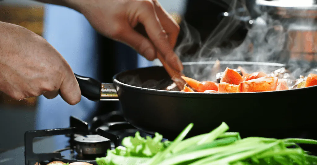 Hands stirring tomatoes in a steaming skillet on a gas burner with spring onions beside the stove