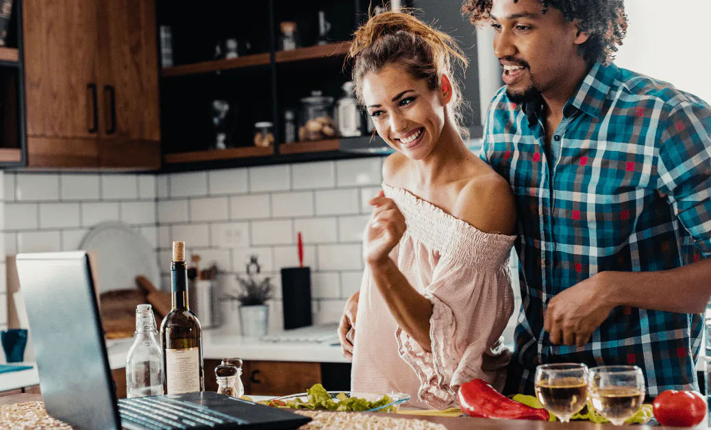 Couple laughing in their kitchen as they cook along with a class on a laptop, wine and vegetables on the counter