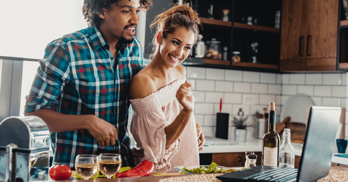 Couple laughing while cooking together at a kitchen counter with a laptop and glasses of wine