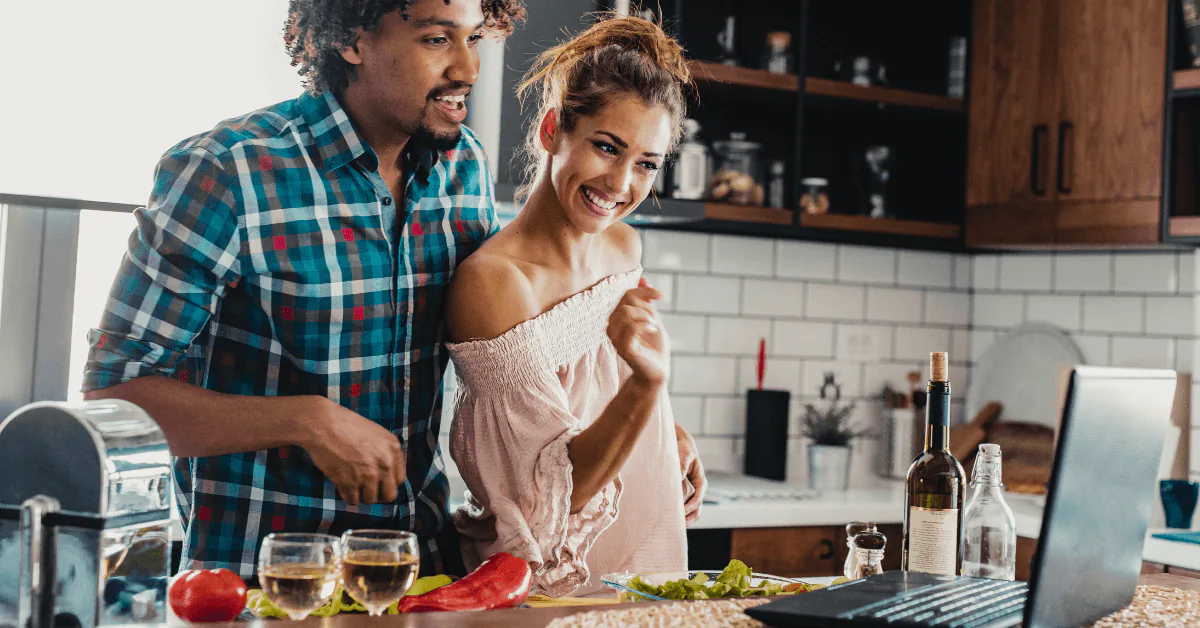 Couple laughing while cooking together at a kitchen counter with a laptop and glasses of wine