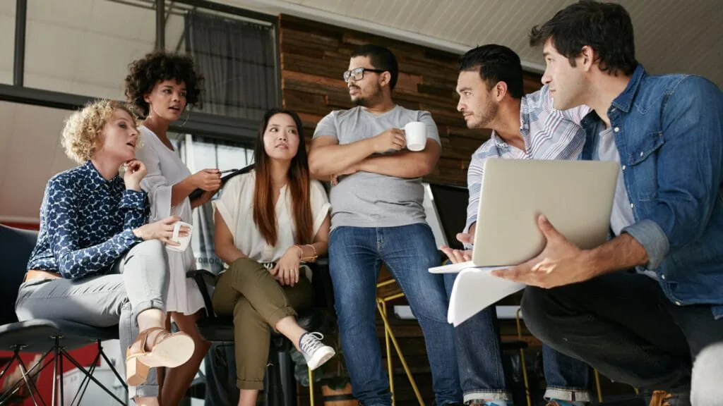 Six coworkers talking casually in an office lounge with a laptop and coffee cups
