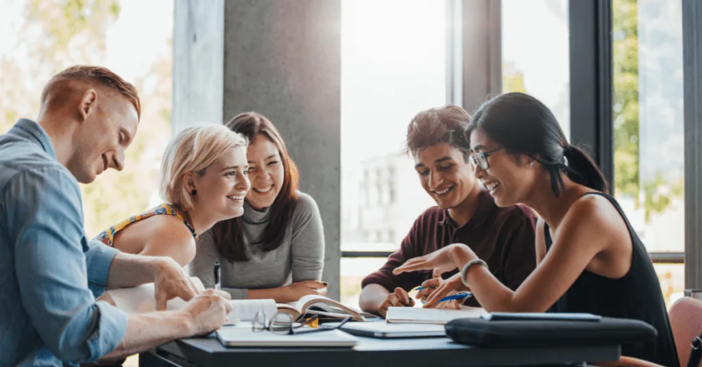 Five coworkers laughing together around a table of notebooks beside a bright window
