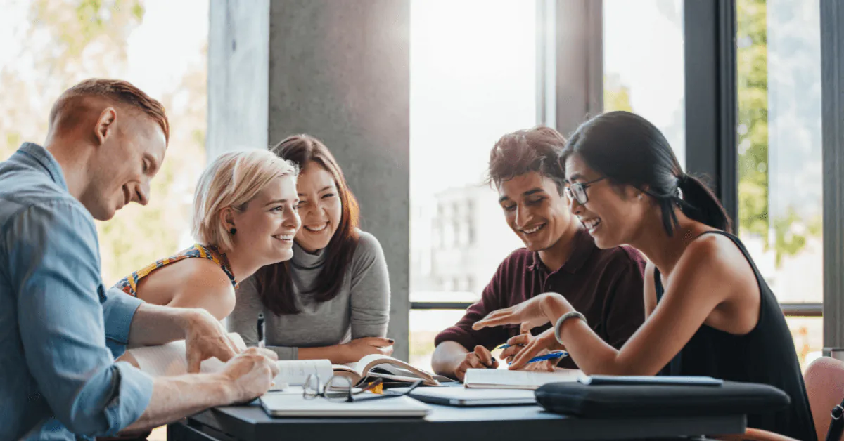 Five colleagues smiling and talking around a table of notebooks beside a bright office window