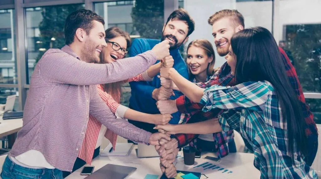Six coworkers laughing as they stack their hands together in the middle of an office