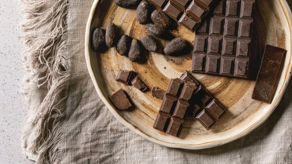 Dark chocolate bars, broken chunks and whole cocoa beans in a wooden bowl on linen cloth