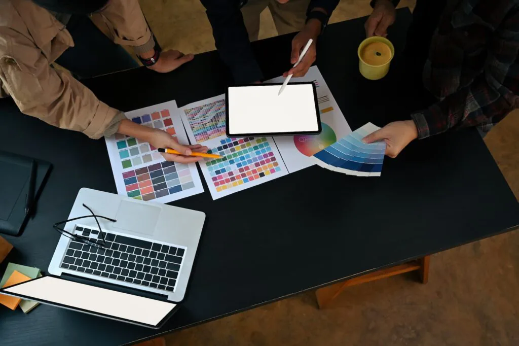 Overhead of a design team comparing color swatch charts, a tablet and a laptop on a dark desk