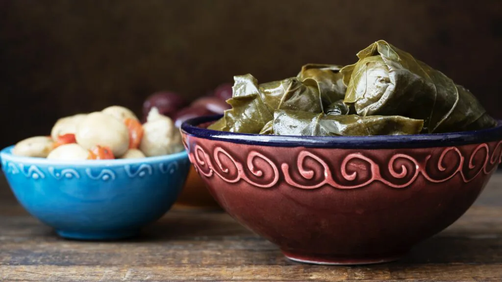 Ceramic bowl of stuffed grape leaves beside a smaller blue bowl of olives and pickled vegetables