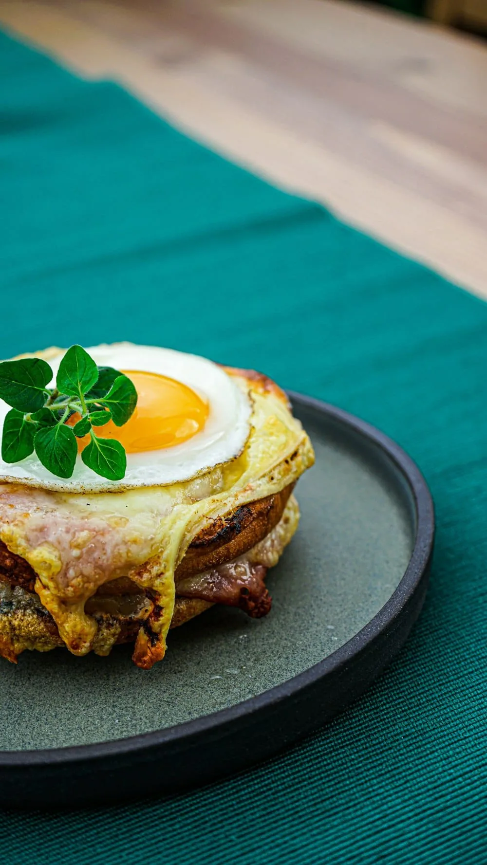 Eggs Benedict topped with a sunny-side-up egg and fresh oregano on a dark round plate
