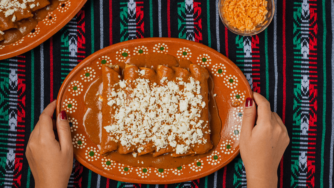 Hands holding a clay plate of enchiladas smothered in mole and topped with crumbled queso fresco