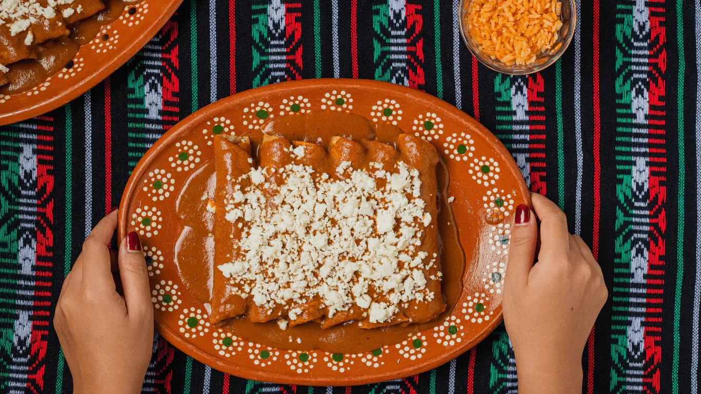 Hands holding a clay plate of enchiladas smothered in mole and topped with crumbled queso fresco
