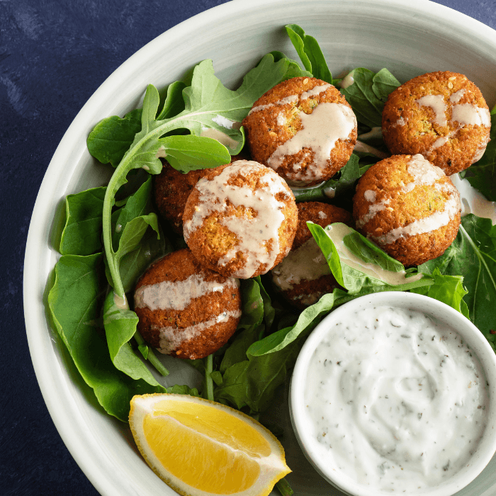 Falafel drizzled with tahini on a bed of greens with herbed yogurt dip and a lemon wedge