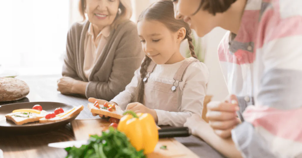 Girl arranging toppings on a wooden board while an older woman and a parent watch in the kitchen